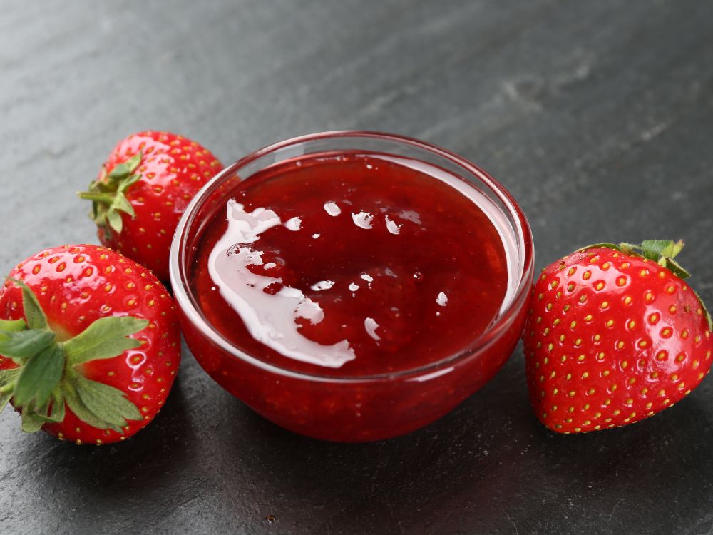 Pote de geleia de morango caseira em recipiente de vidro, acompanhado de morangos frescos sobre superfície escura.